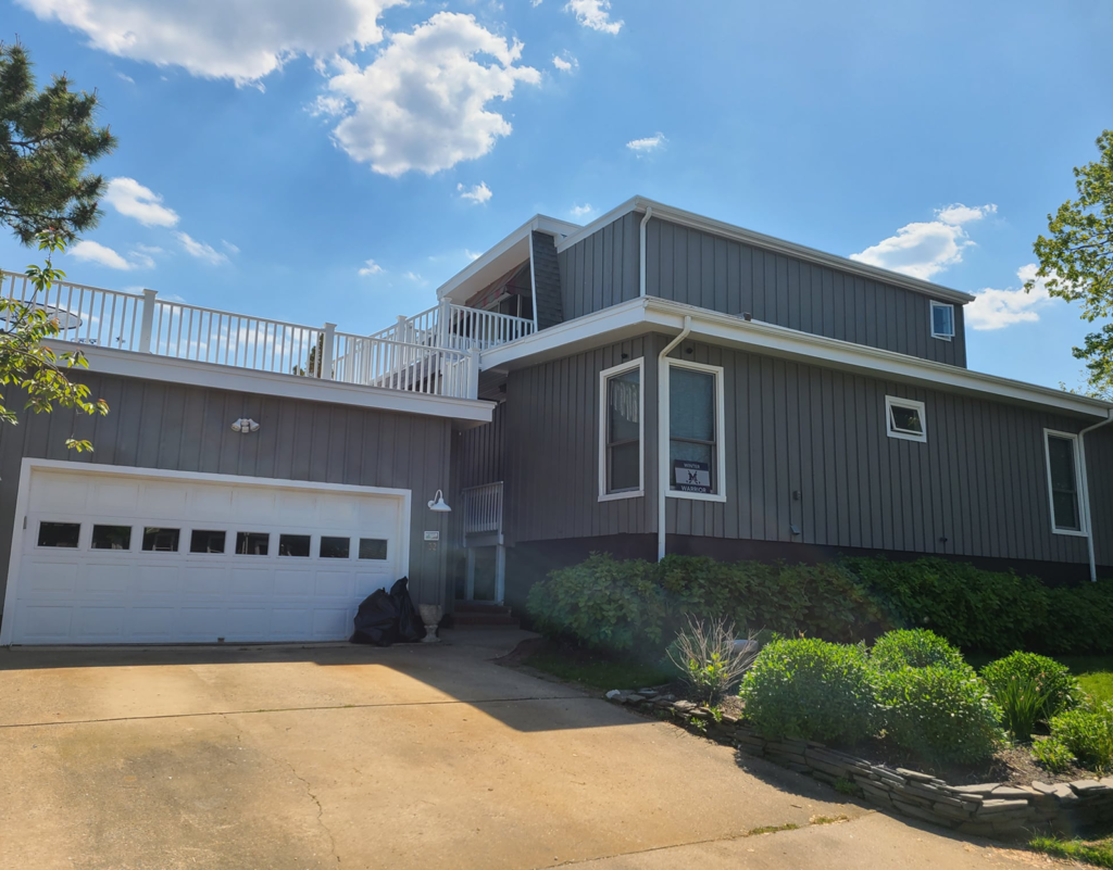 Exterior view of a modern residential home with gray siding, white trim, and a spacious balcony, showcasing a well-maintained driveway and landscaped garden, reflecting J.A. Painting & Decorations' high-quality exterior painting services.